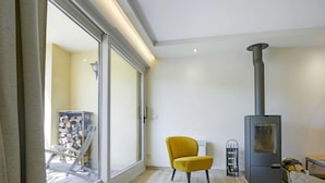 Ein modernes Wohnzimmer mit einer großen Schiebeglasdoor, einem gelben Sessel, einem schwarzen Kaminofen und einer weißen Heizkörper.