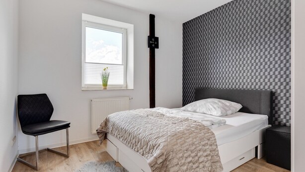 Une chambre à coucher comprenant un lit, une chaise, un radiateur, une fenêtre et un mur orné d’un motif à carreaux.