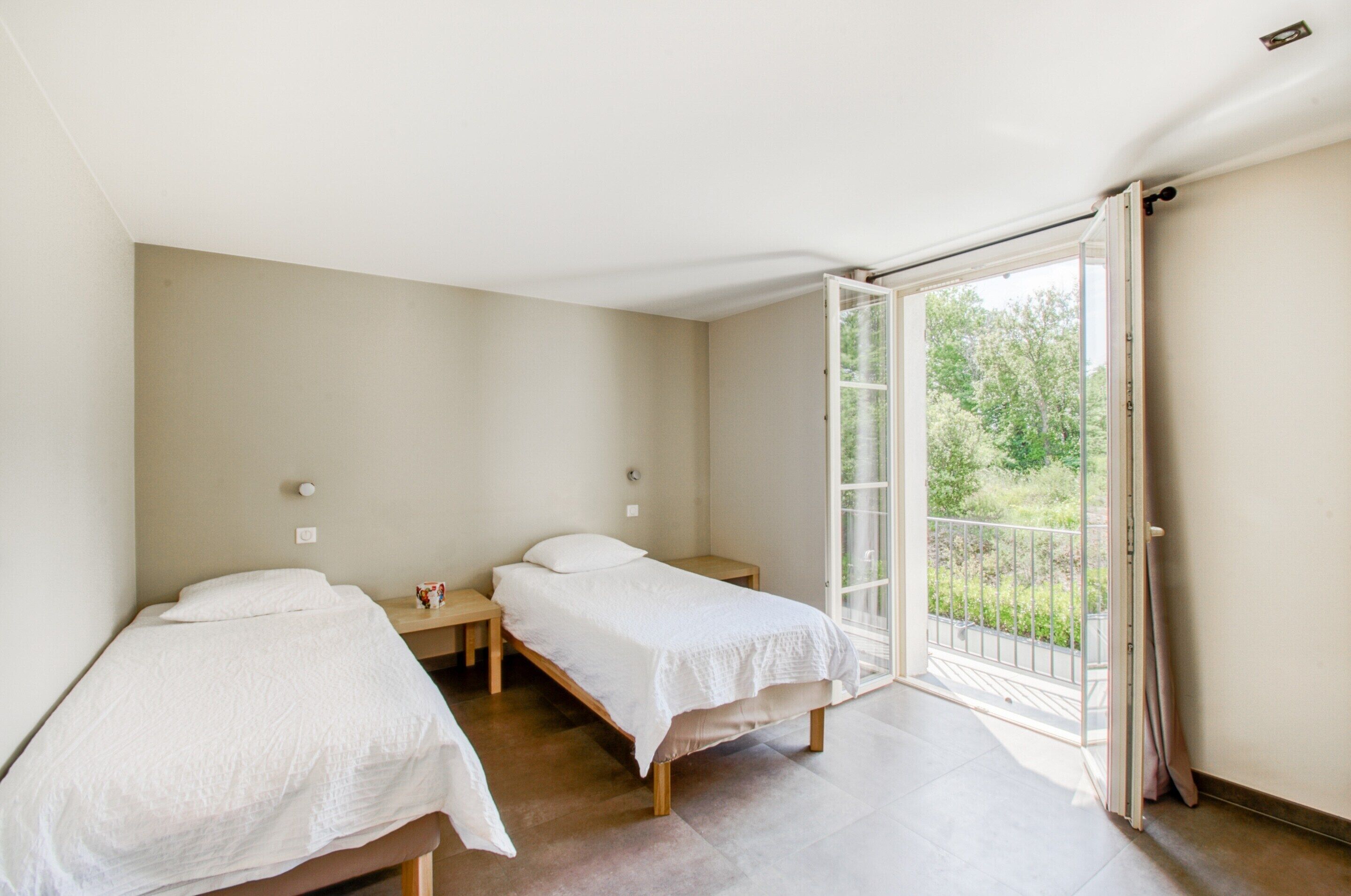A room with two single beds, a small table, and a sliding glass door leading to an outdoor balcony.