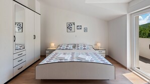 A modern bedroom with a large bed, bedside tables, a wardrobe, and a view of greenery through a large window. - Bonaventura - Countryside Villa near Split with Private Pool (Muc)