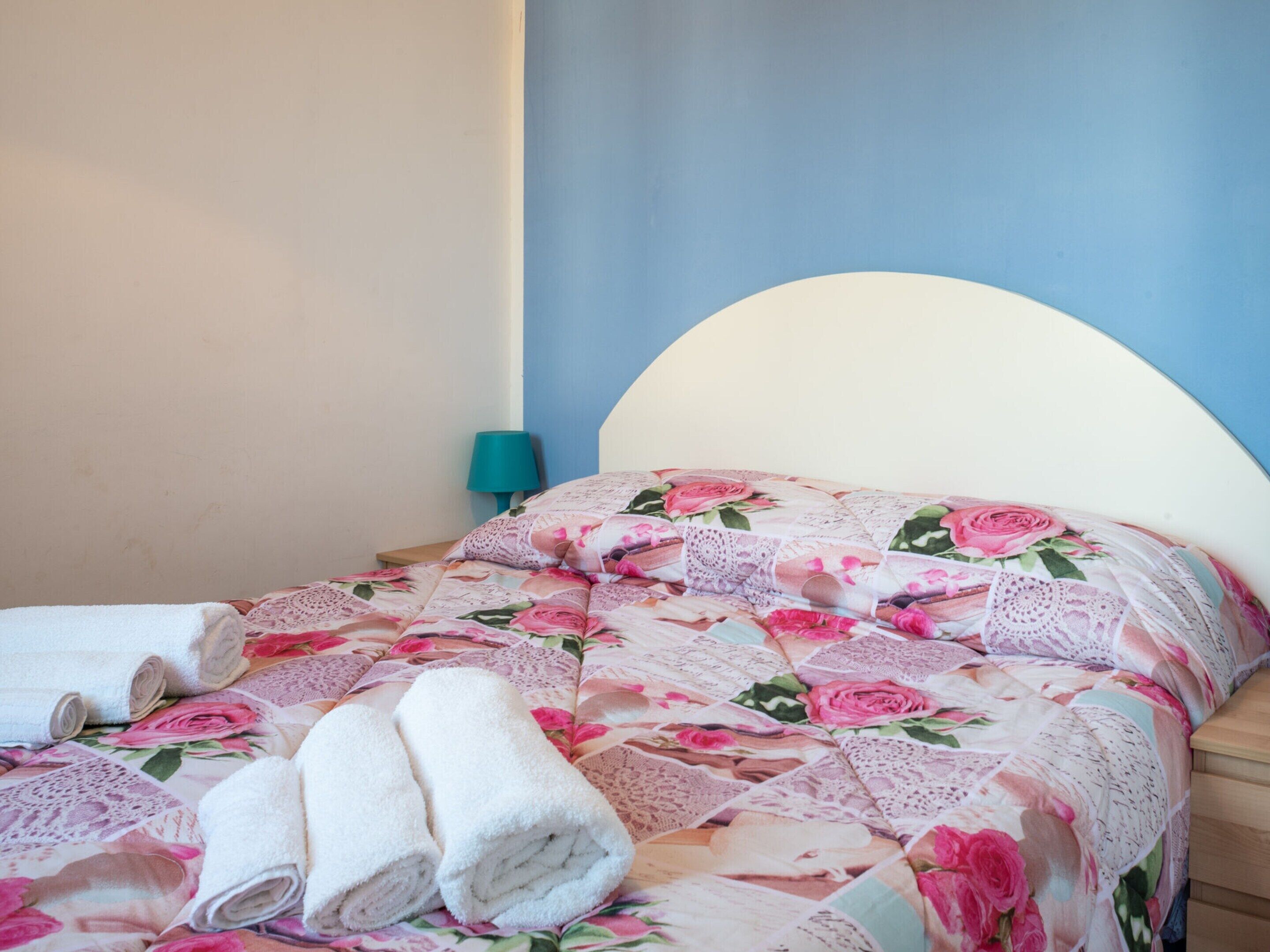 A bed with a floral bedspread, two rolled towels, and a teal nightstand with a lamp.