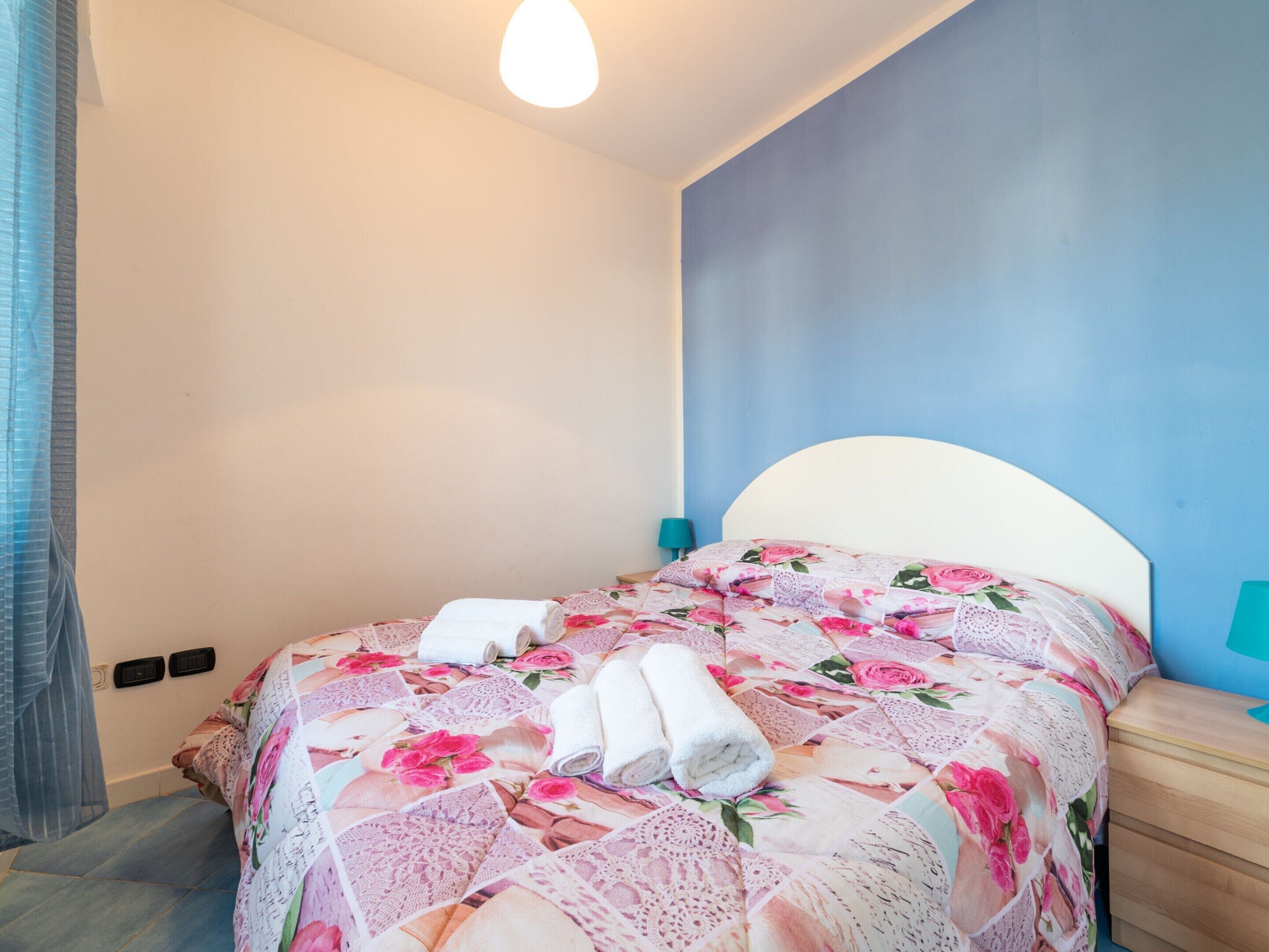 A neatly made bed with floral bedding, a bedside lamp, and a wall with a blue accent.