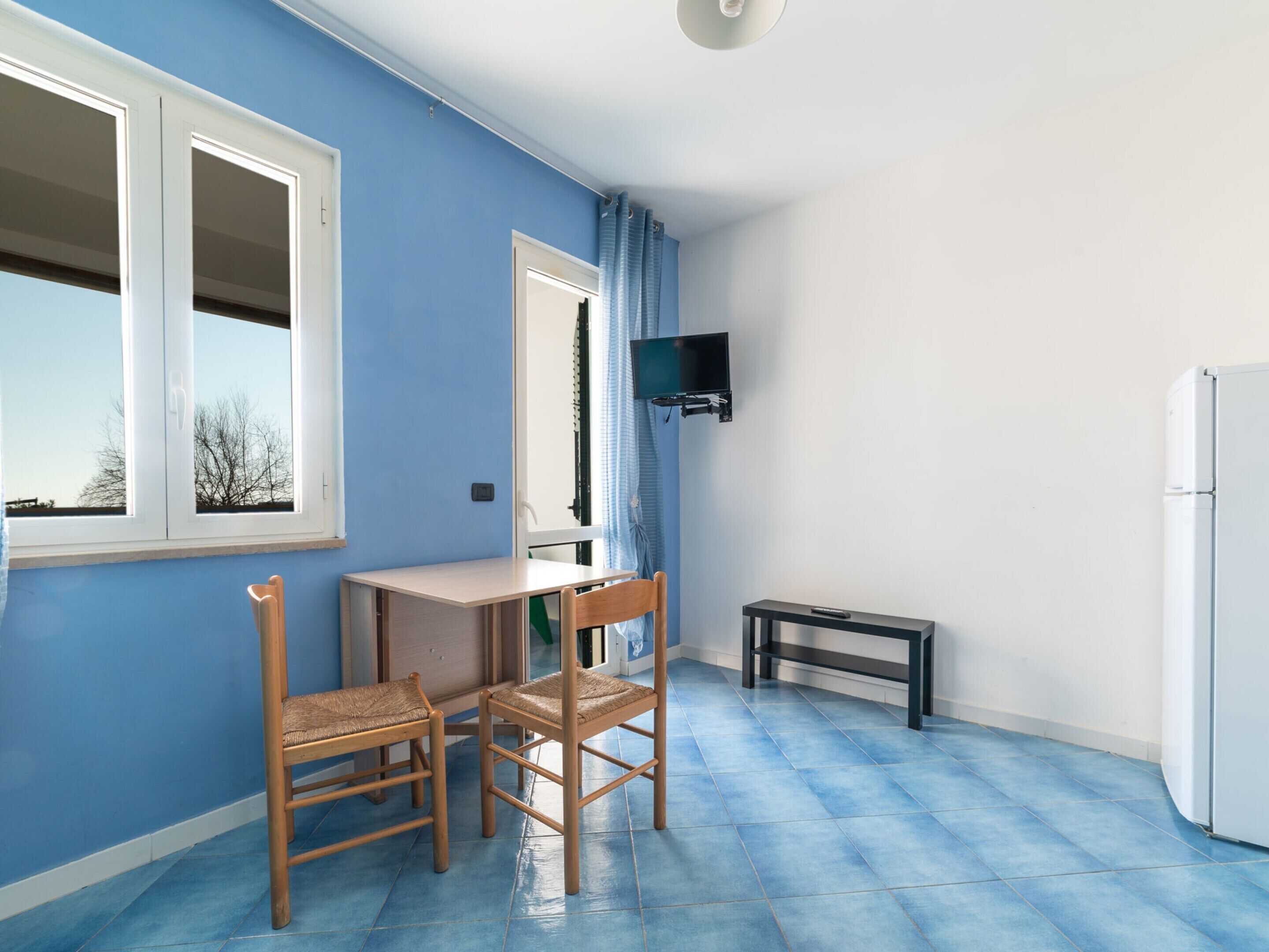 Une pièce avec un mur bleu, une porte blanche, une petite table, deux chaises, un téléviseur fixé au mur et un réfrigérateur blanc.