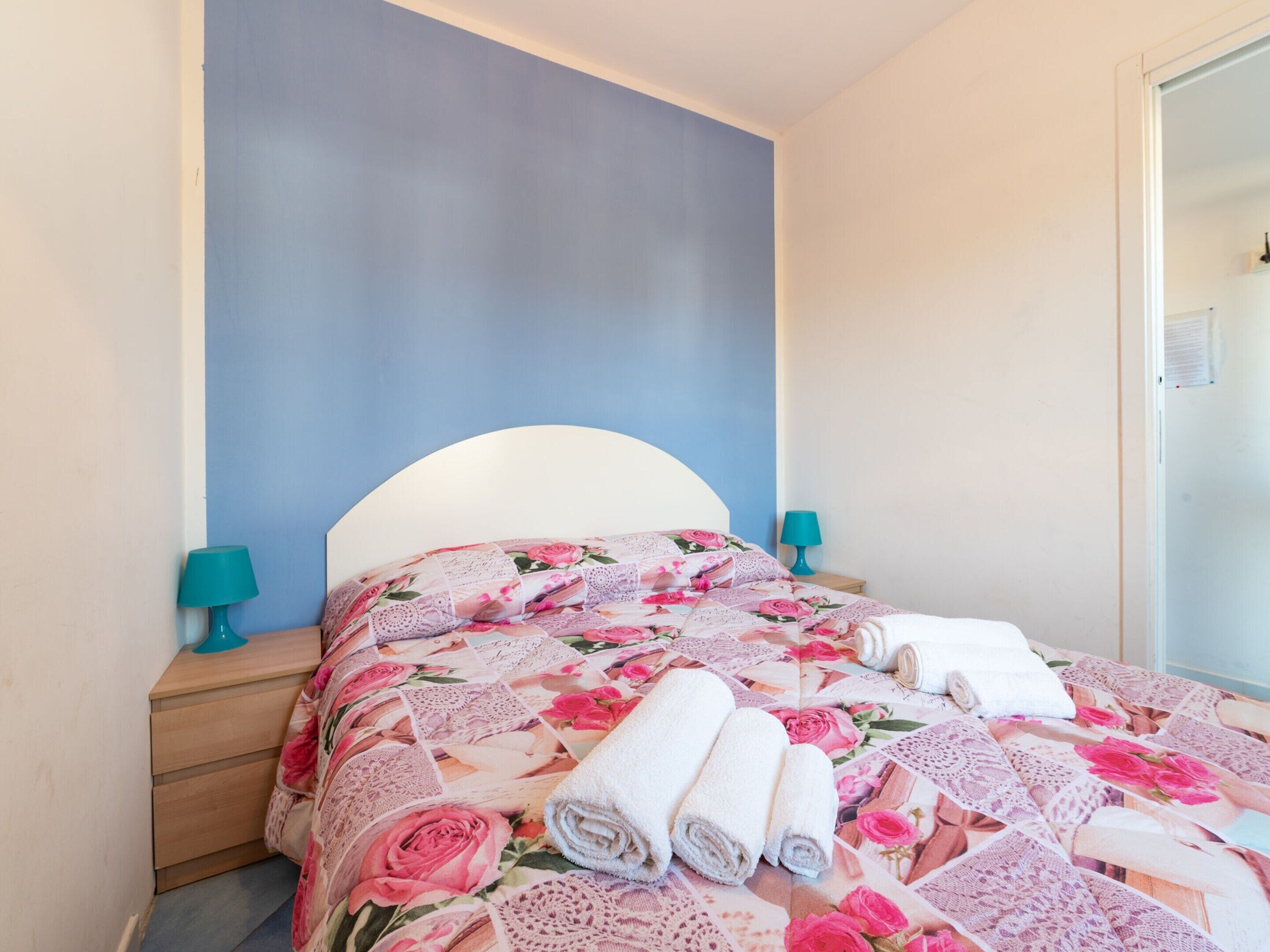 A neatly made bed with floral bedding, two bedside lamps, and rolled towels.