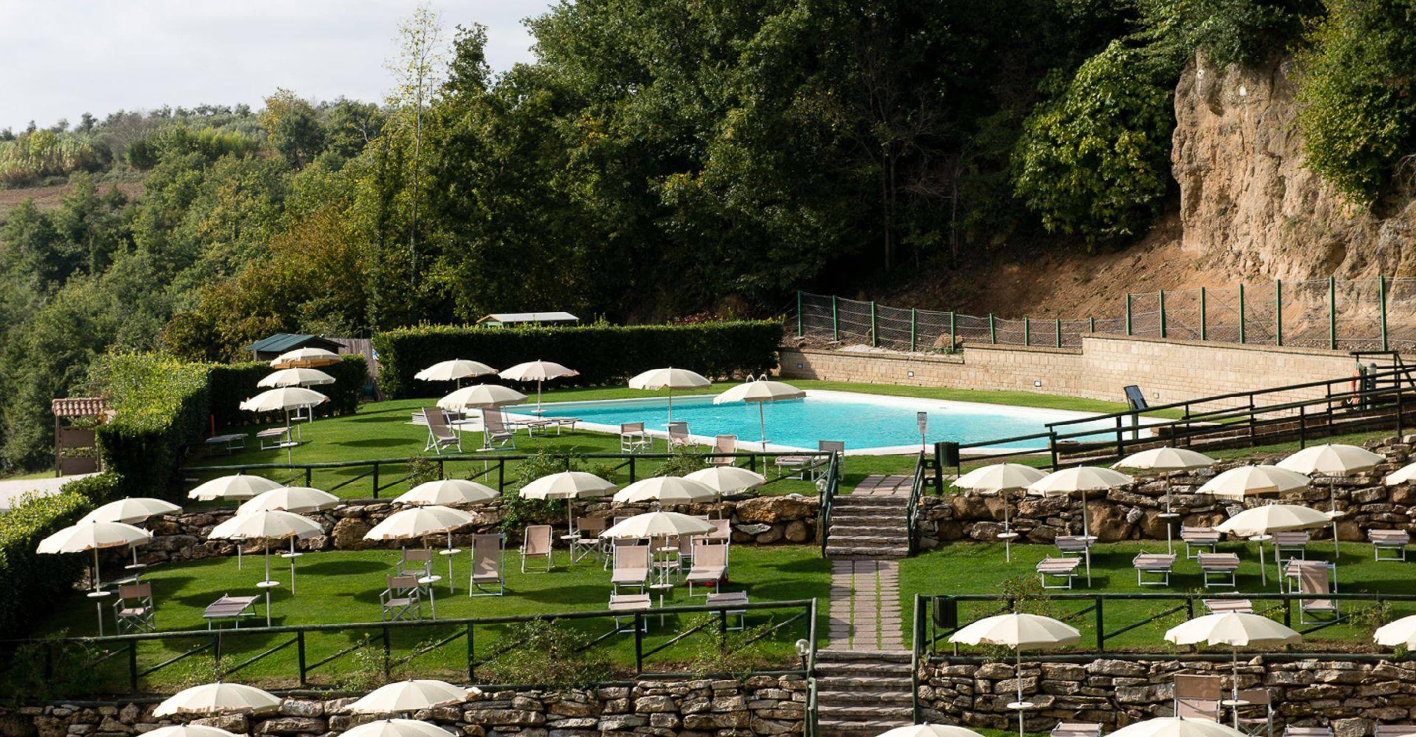 Ein Poolbereich mit mehreren Liegestühlen und Sonnenschirmen, umgeben von Steinmauern und viel Grün.