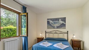 Une chambre à coucher avec un lit, deux tables de chevet avec des lampes, un radiateur, une fenêtre avec des rideaux et un tableau accroché au mur.