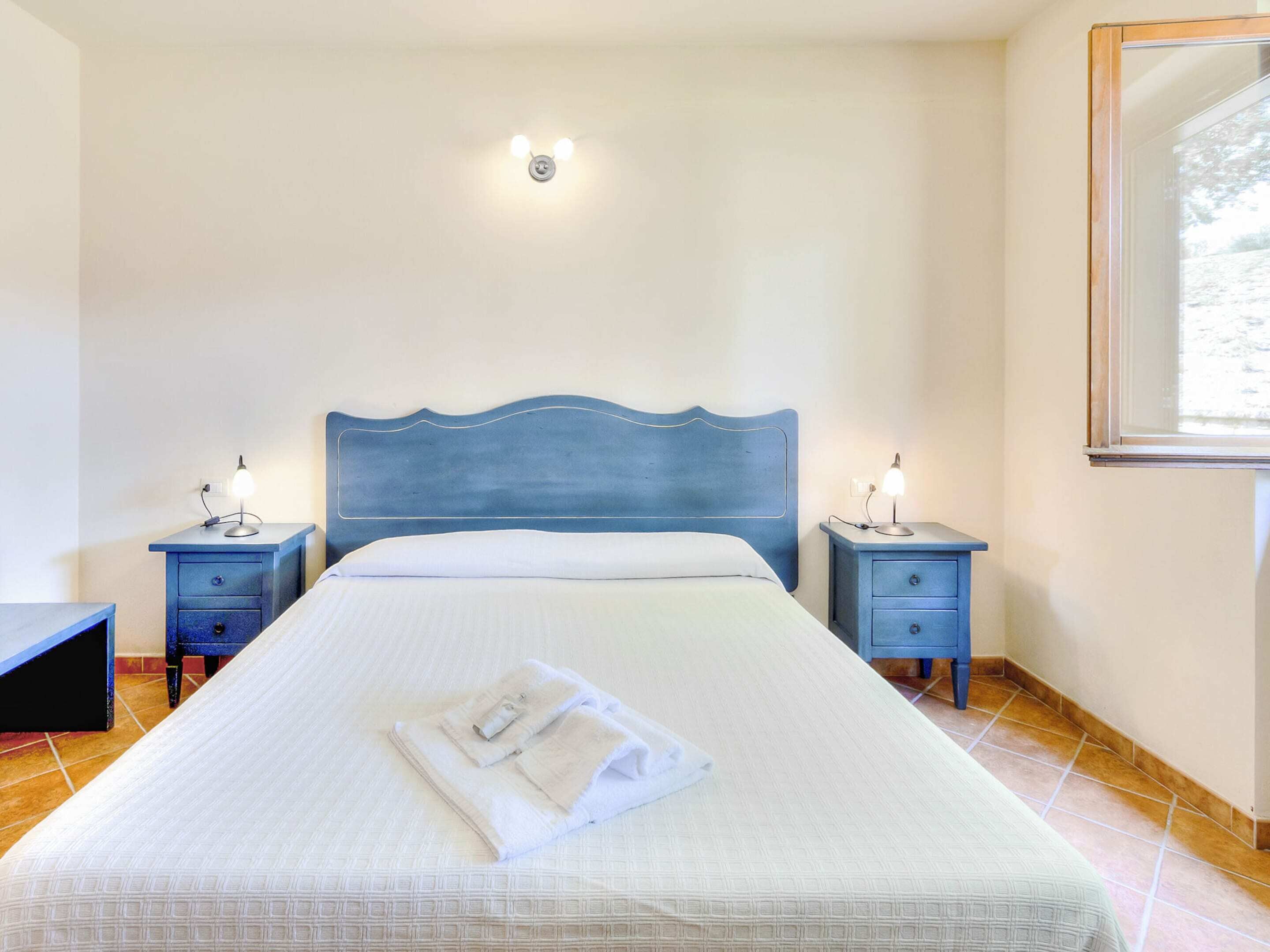 A neatly made bed with a blue headboard, two bedside tables with lamps, and a window with a view of trees.