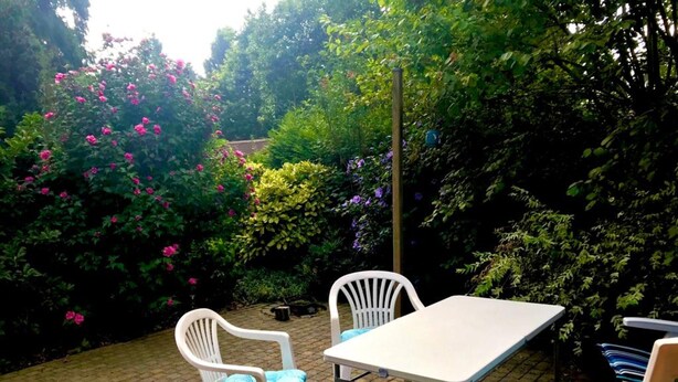 Um pátio externo com uma mesa e duas cadeiras, cercado por vegetação exuberante e plantas floridas.