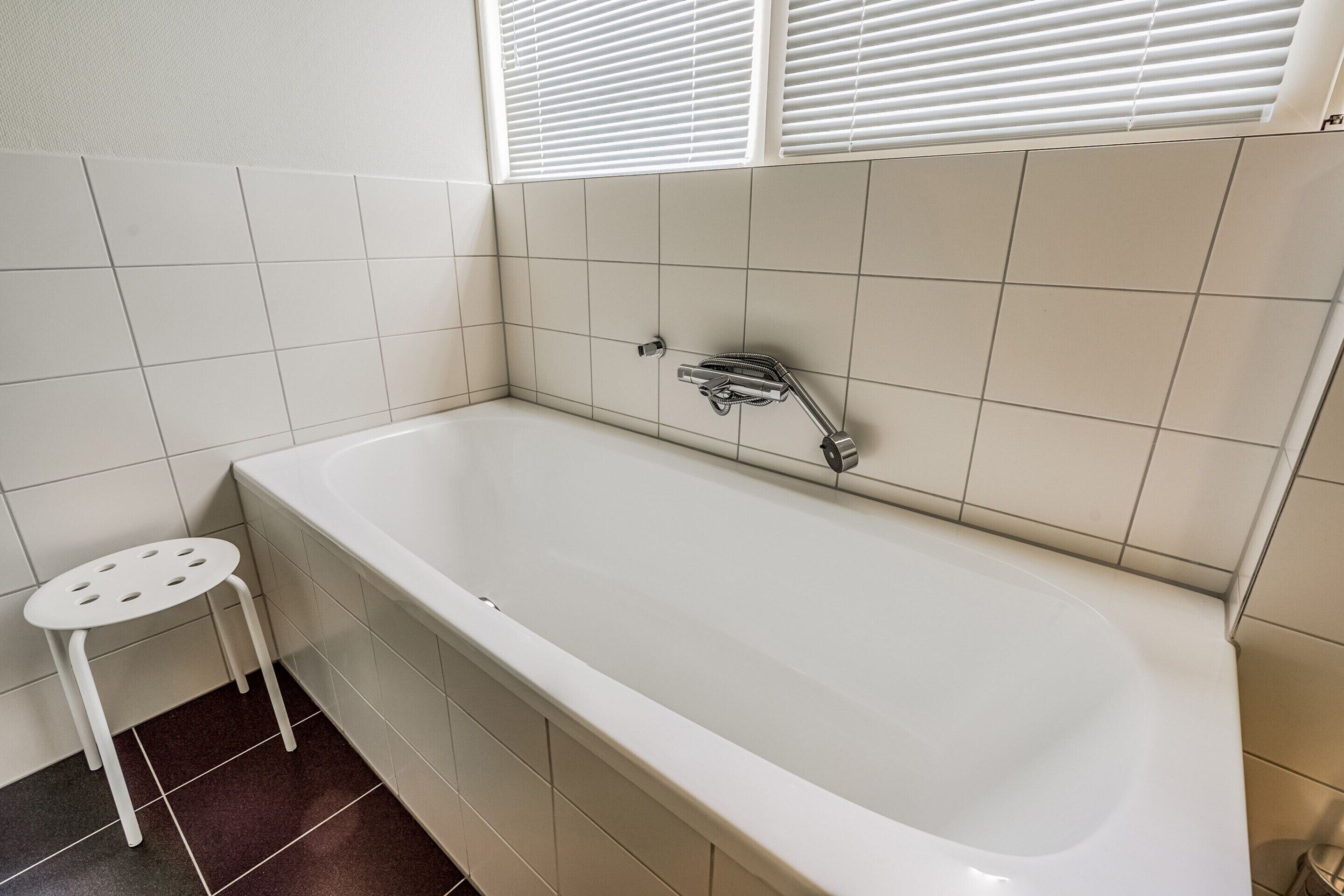 A bathroom with a white bathtub, a small white stool, and a window with blinds.