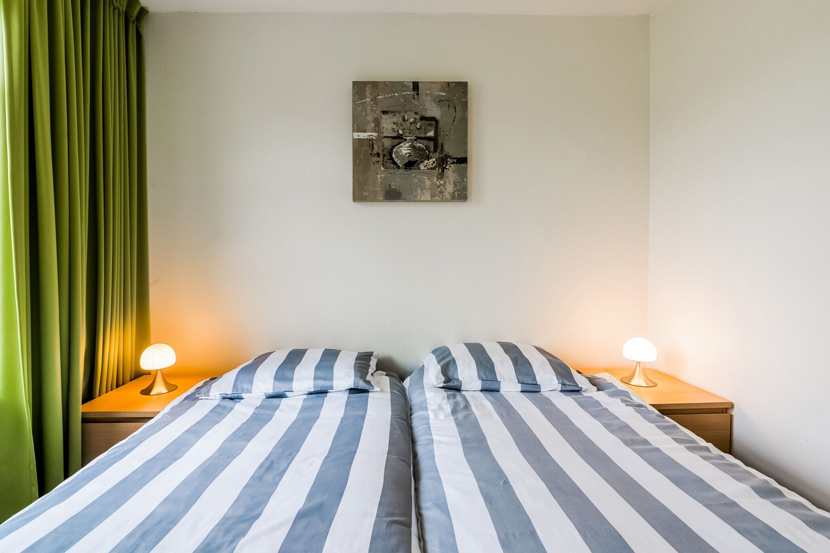 Cama doble con ropa de cama a rayas, dos lámparas de noche, una cortina verde y una obra de arte abstracta colgada en la pared.