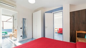 A bedroom with a red bedspread, a wooden chair, a wardrobe, and a view of a balcony with a red chair. - Belvilla by OYO Apartment in Policastro Bussentino (Santa Marina)