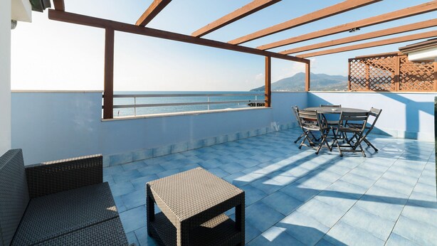 A rooftop terrace with a table and chairs, a wicker sofa, and a view of the sea. - Belvilla by OYO Apartment in Policastro Bussentino (Santa Marina)