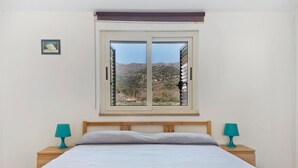 Ένα υπνοδωμάτιο με ένα κρεβάτι, δύο κομοδίνα, ένα παράθυρο με θέα σε λόφους και έναν μικρό πίνακα στον τοίχο.