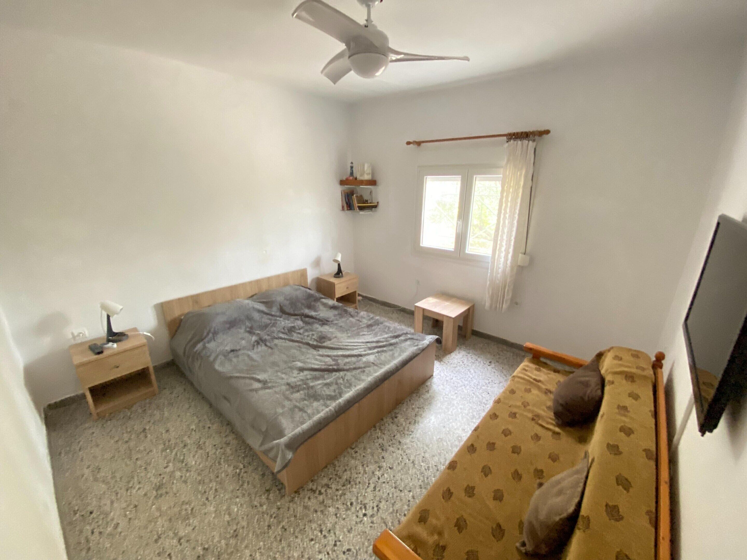 Un dormitorio con una cama, una mesita de noche, un taburete pequeño, un televisor y un ventilador de techo.