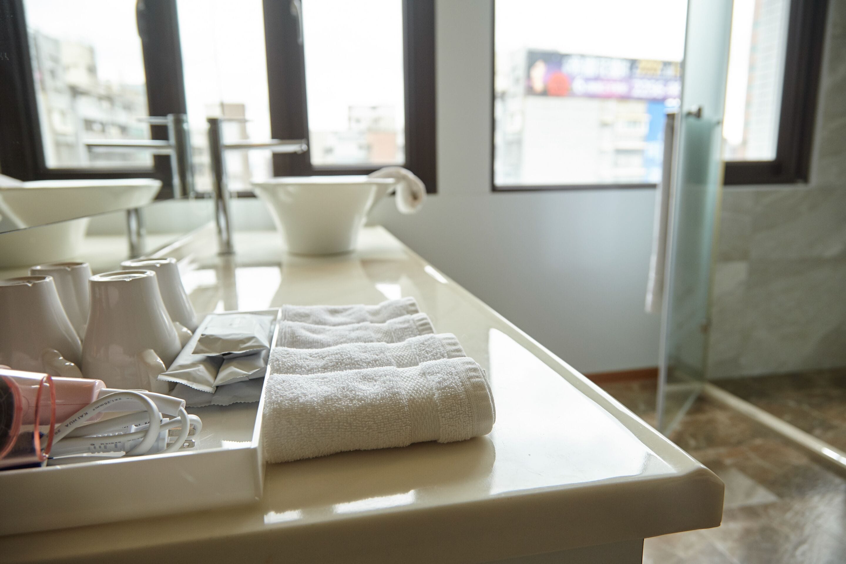 A bathroom with a white countertop, a sink, and a window with a view of buildings.