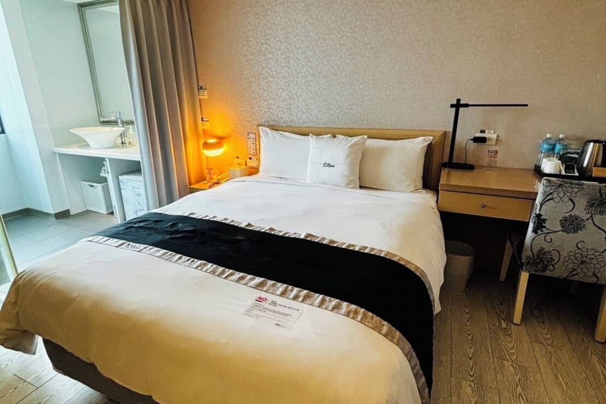 A hotel room with a bed, bedside table, lamp, and a desk with a chair.