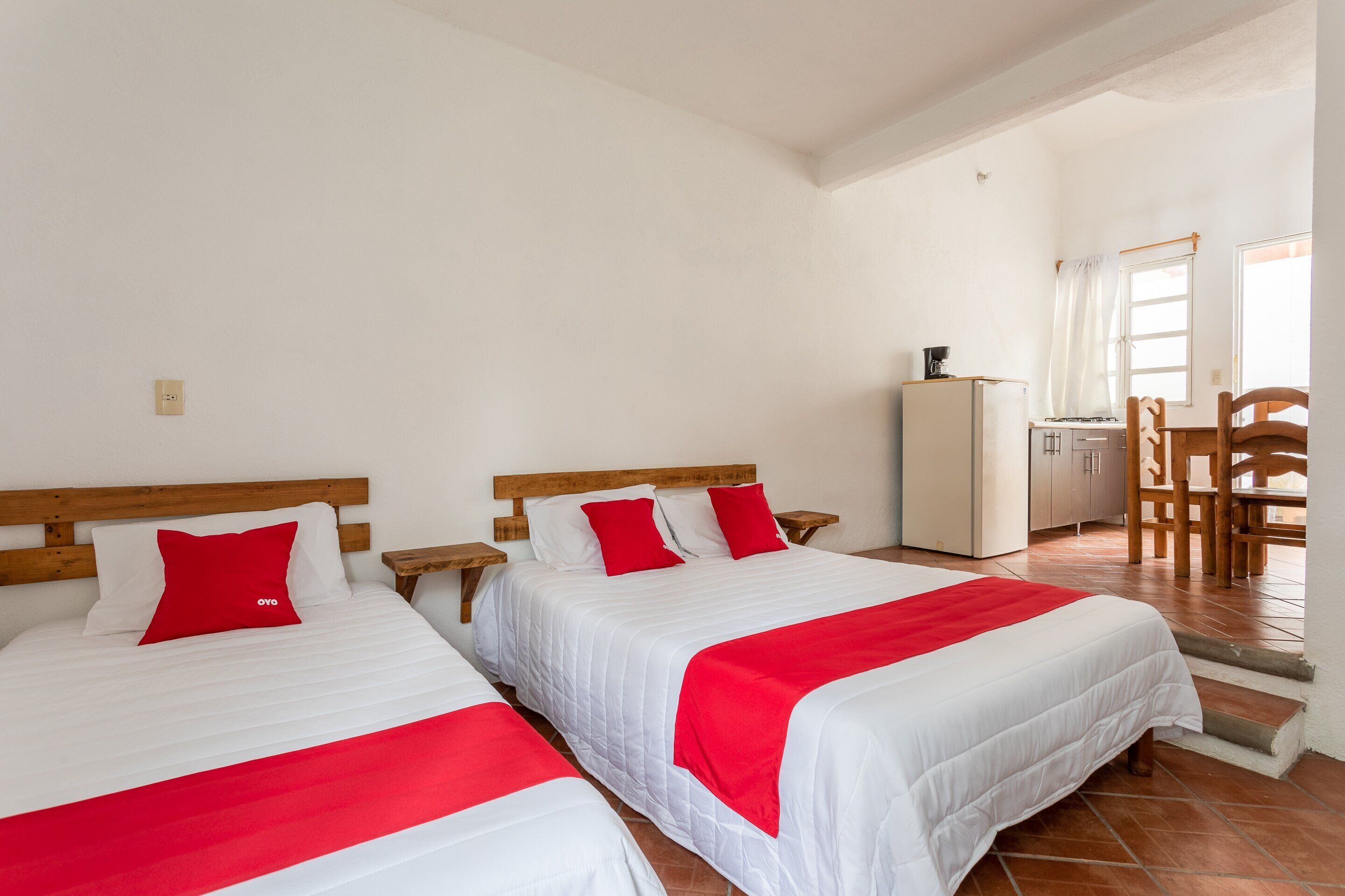A hotel room with two beds, each with white bedding and red accents, a wooden headboard, and a small kitchenette in the background.