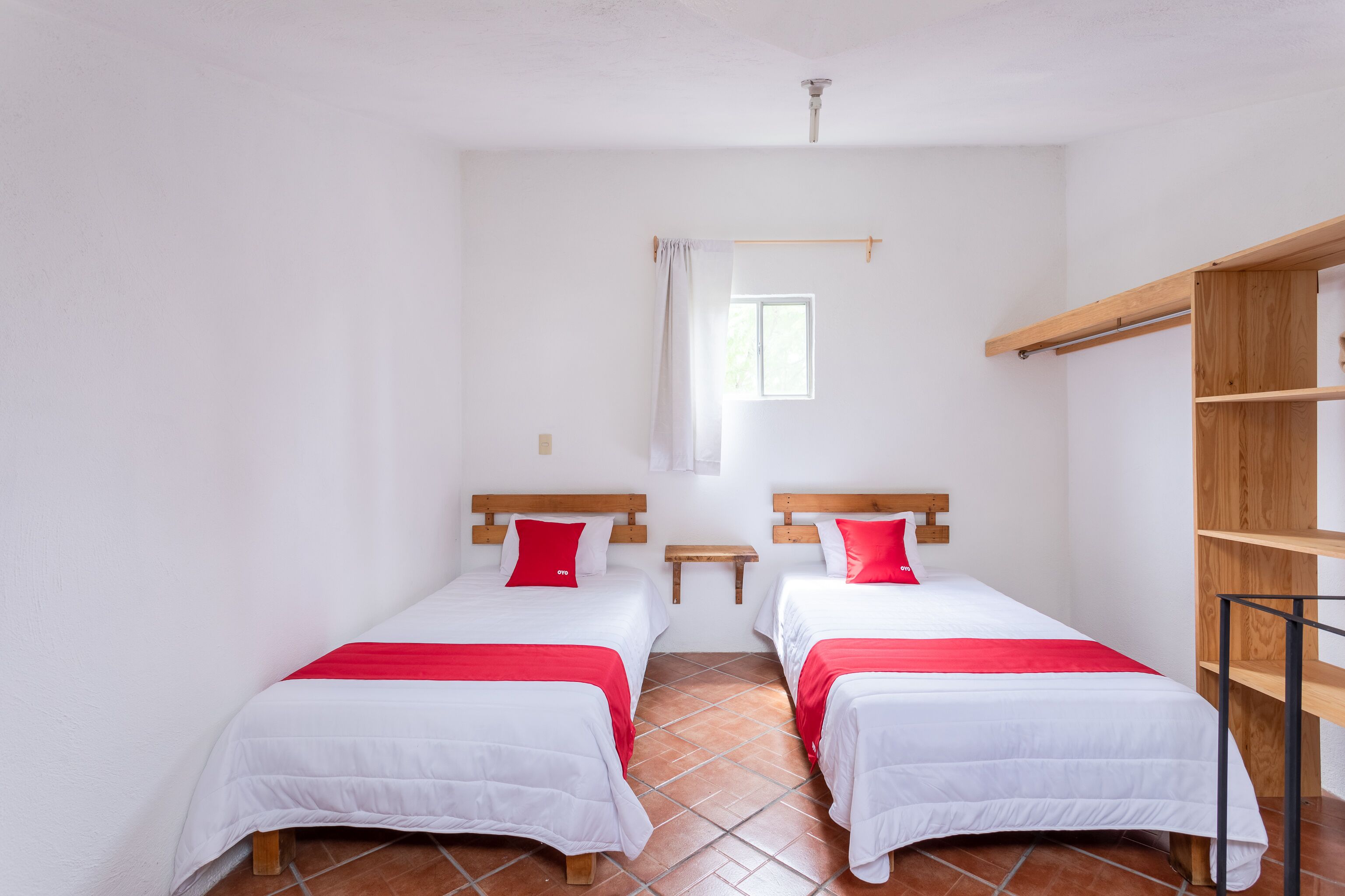 A room with two beds, each with a red pillow and white bedding, a small window, and a wooden shelf unit.