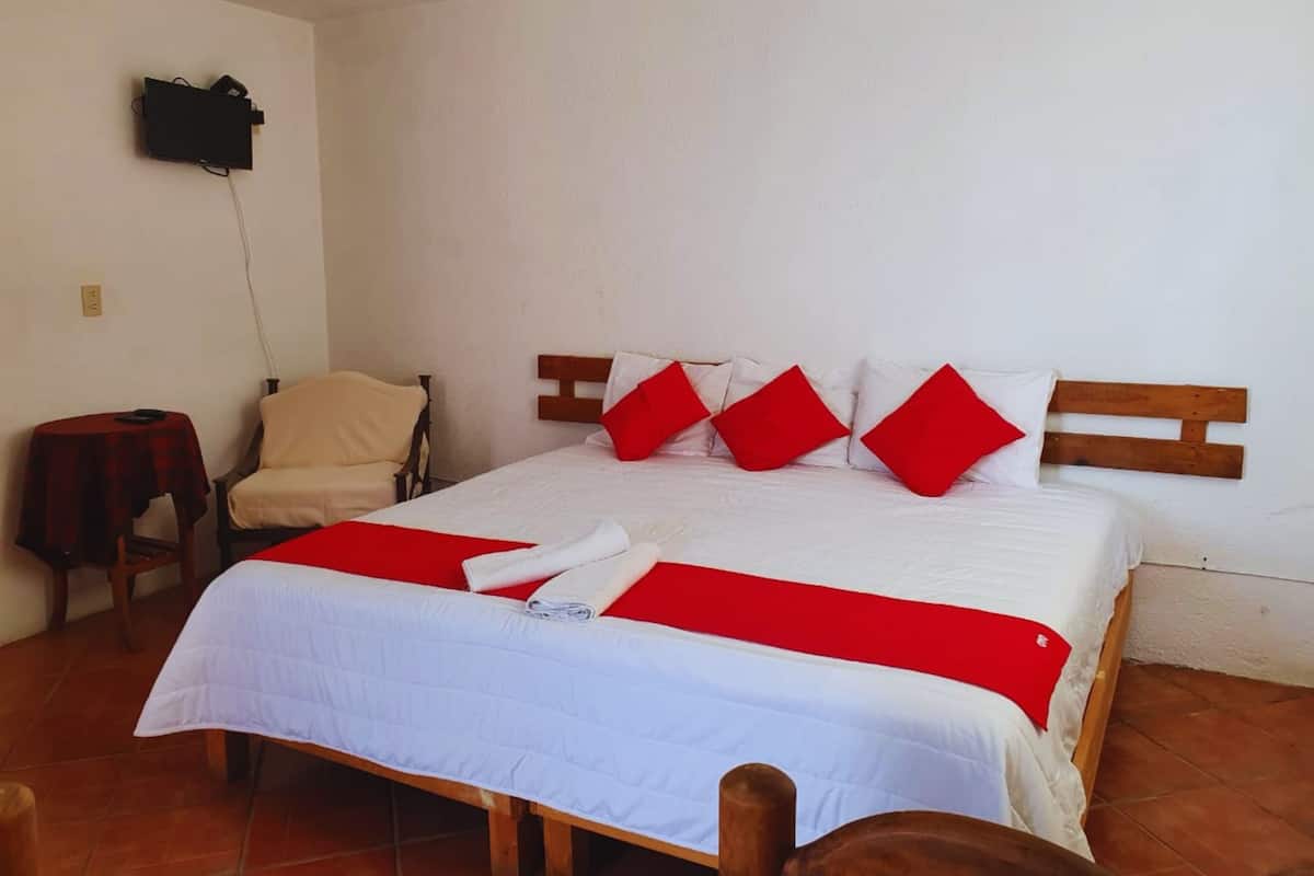 Un lit bien fait, recouvert d’une couvre-lit rouge et blanche, avec une tête de lit en bois et des oreillers rouges. Une petite table recouverte d’une nappe rouge et une chaise se trouvent à gauche.