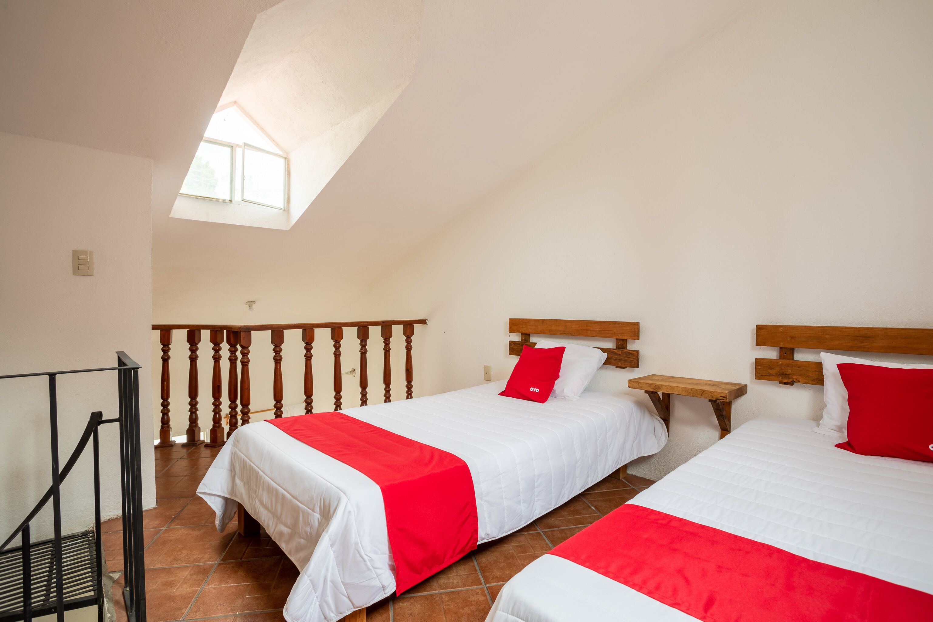 Habitación con dos camas, cada una con ropa blanca y detalles rojos, un cabecero de madera y una mesita de noche de madera.