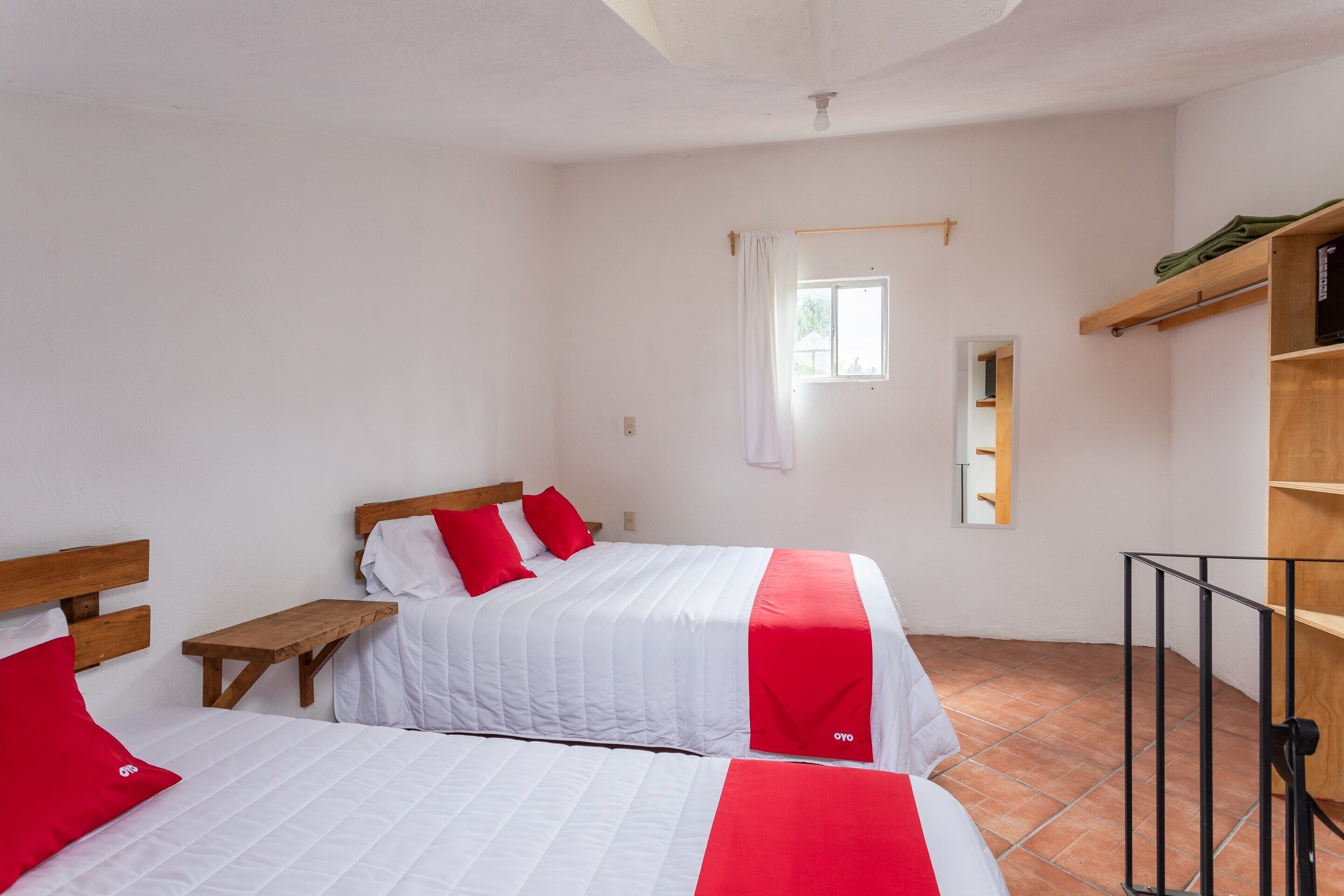 Una habitación con dos camas, cada una con ropa de cama roja y blanca, un banco de madera, una mesita pequeña y un estante de madera con un espejo.