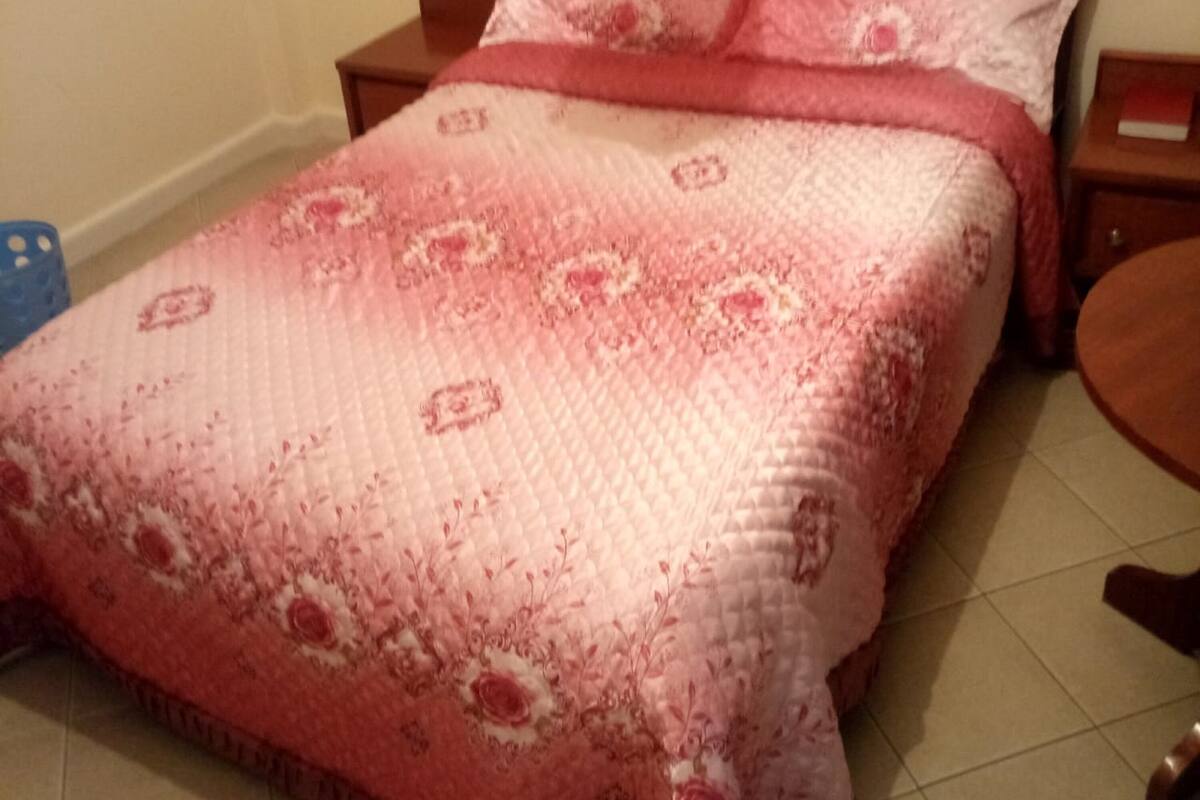 A bed with a pink floral bedspread and matching pillows, a wooden nightstand, and a blue chair.