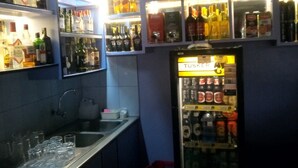 Una zona de bar con estantes llenos de botellas de licores, un refrigerador repleto de bebidas y un mostrador con vasos y una caja registradora.