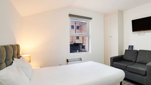 A bedroom with a bed, a window with a view of a brick building, a grey sofa, and a flat-screen TV mounted on the wall. - Modern Studio in London Near Wimbledon Park (London)