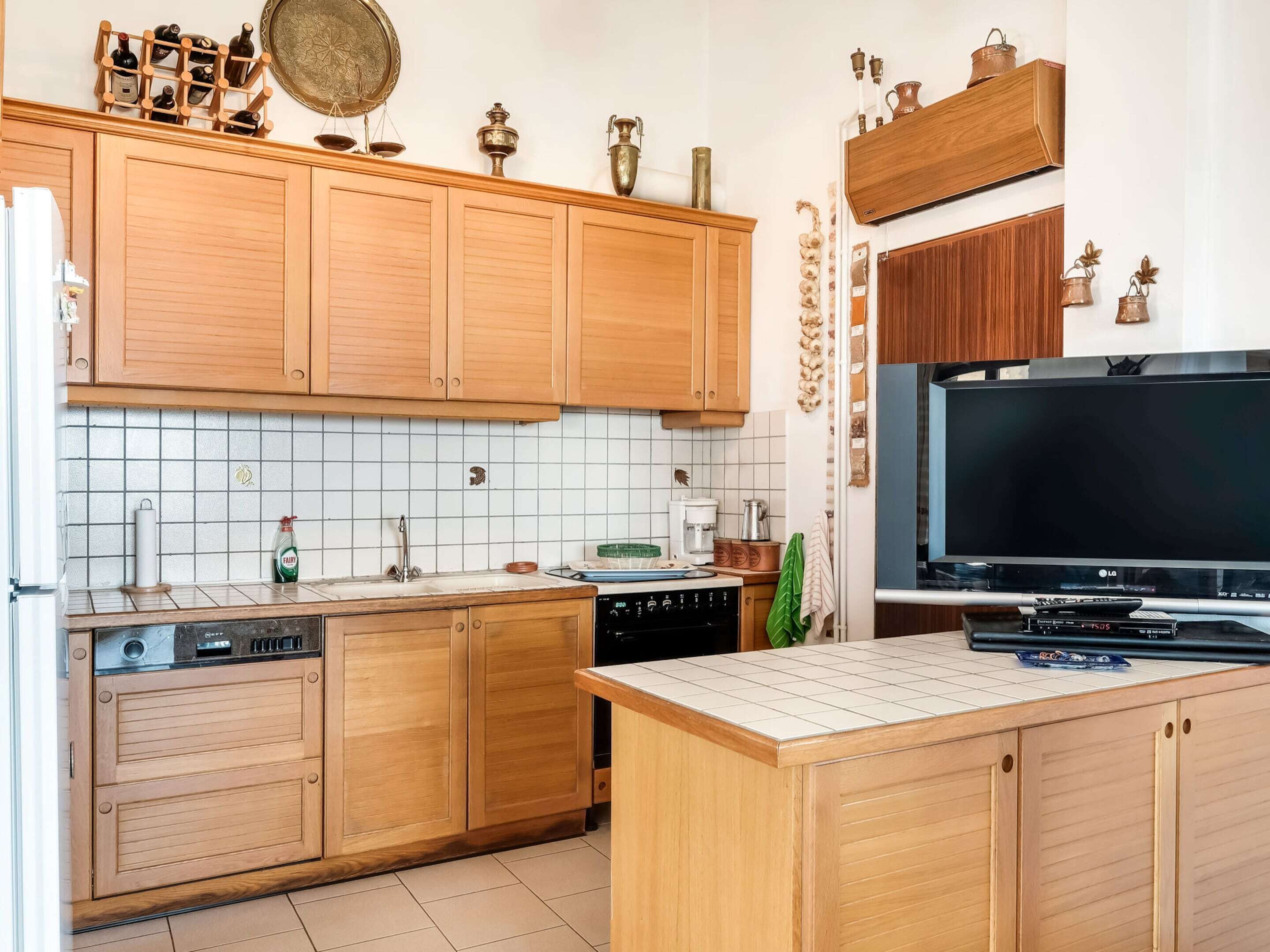 A kitchen with wooden cabinets, a tiled backsplash, a built-in dishwasher, a coffee maker, and a flat-screen TV.