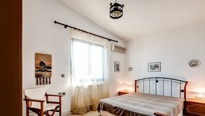 A bedroom with a bed, a chair, a bedside table, a hanging light fixture, and a window with sheer curtains. - Holiday Villa With sea View in Paralia Avlidas (Chalcis)