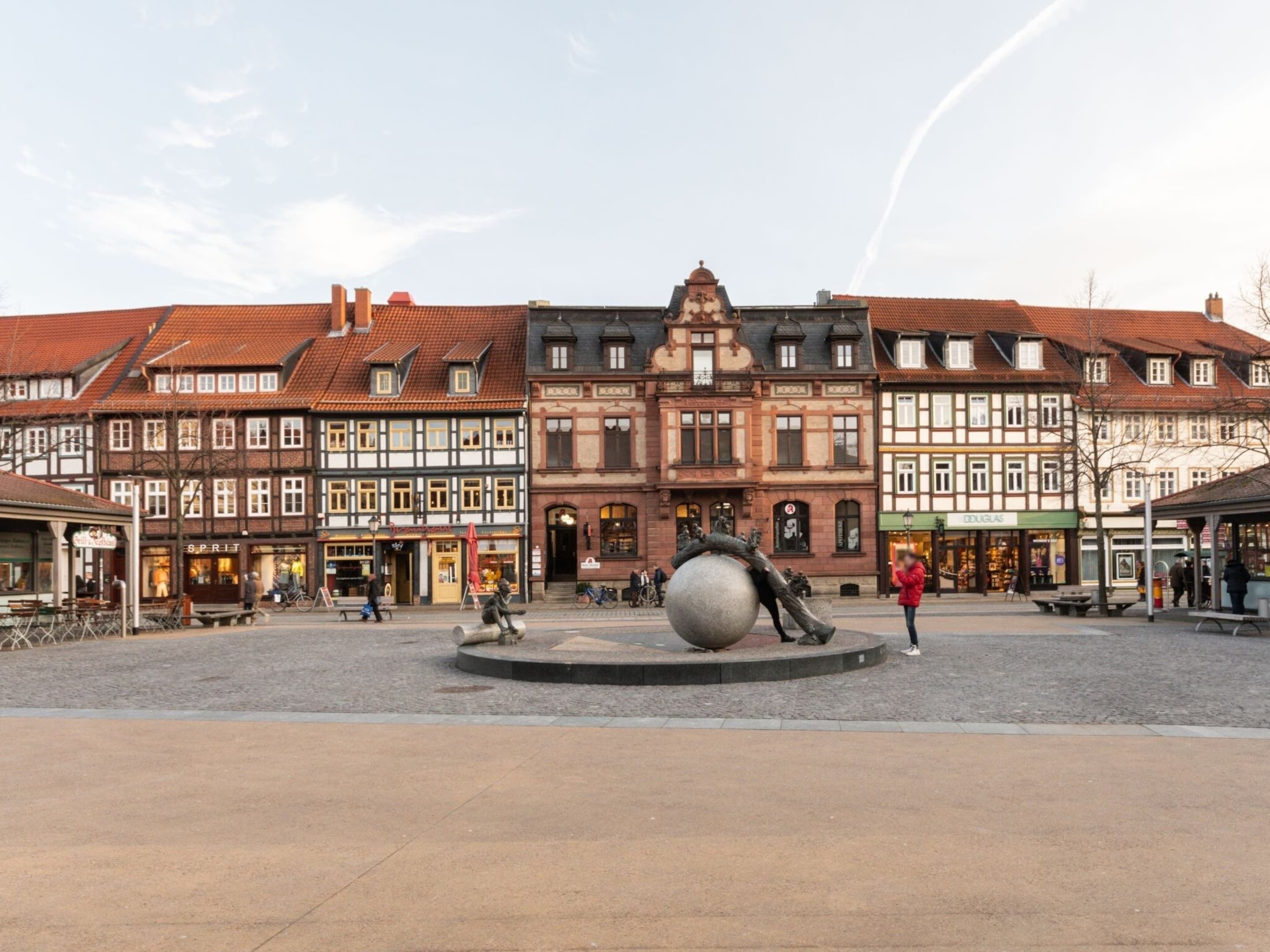 A public square with historic buildings, a large sculpture, and a few people.