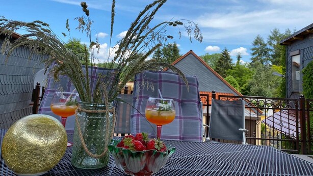 An outdoor patio with a table set for two, featuring a glass jar with a plant, a bowl of strawberries, and two glasses of orange drink.