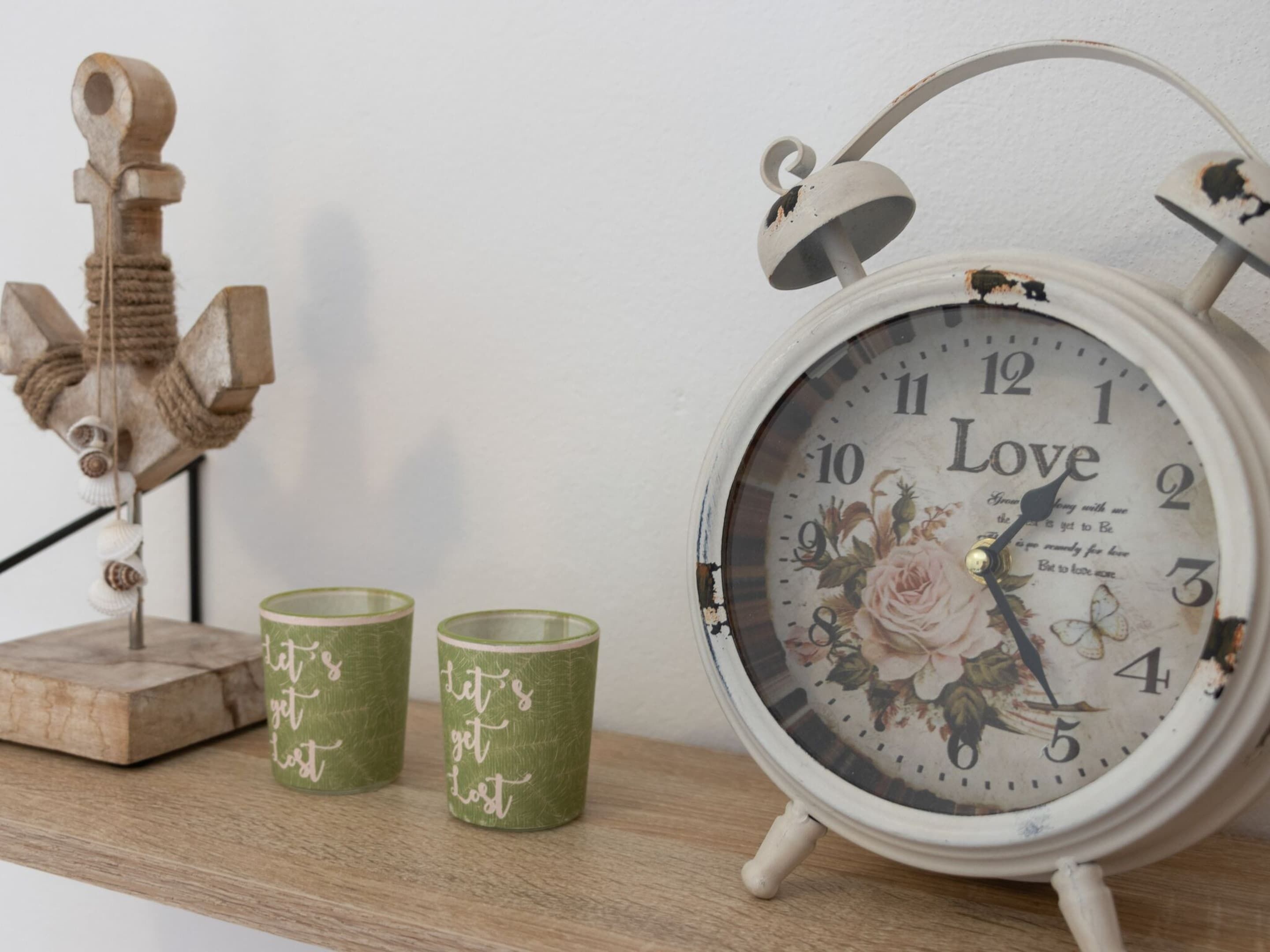 Un reloj de estilo vintage con diseños florales y la palabra 'Love' en su esfera, colocado sobre un estante de madera junto a dos candelabros verdes con la frase 'let's get lost'.