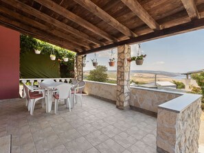 Une terrasse couverte avec des piliers en pierre, des meubles d’extérieur blancs, des jardinières suspendues et une vue sur le paysage.