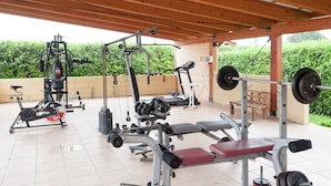 Ein Outdoor-Fitnessbereich mit verschiedenen Trainingsgeräten, darunter ein Laufband, eine Gewichtbank und Kurzhanteln.