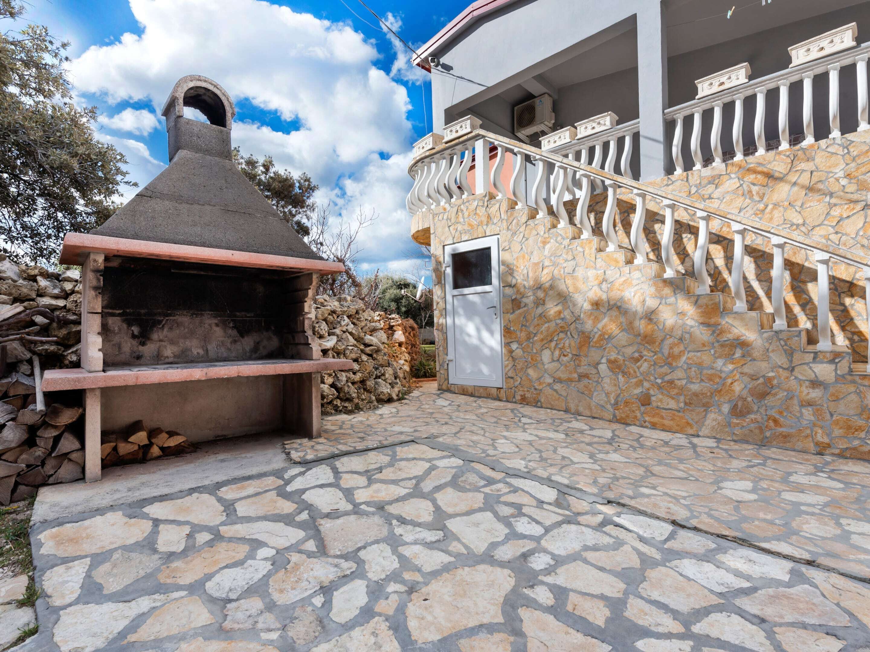 Une terrasse extérieure en pierre, avec un barbecue en pierre, un mur en pierre et un escalier doté de balustrades blanches.