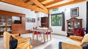A cozy room with wooden beams, a dining table with chairs, a sofa, a cabinet with dishes, and a window with curtains. - Homes in the Lagoons of Santa Giusta (Santa Giusta)