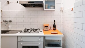 Une cuisine compacte aux murs recouverts de carreaux blancs, avec une cuisinière à gaz, un four encastré, un four à micro-ondes et un petit plan de travail sur lequel se trouvent une bouteille et une horloge.