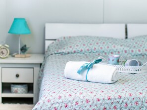 A neatly made bed with a floral duvet, a white bedside table with a lamp, and a small shelf with a clock and decorative items. - Secluded Holiday Home in Pašman near Sea (Pasman)