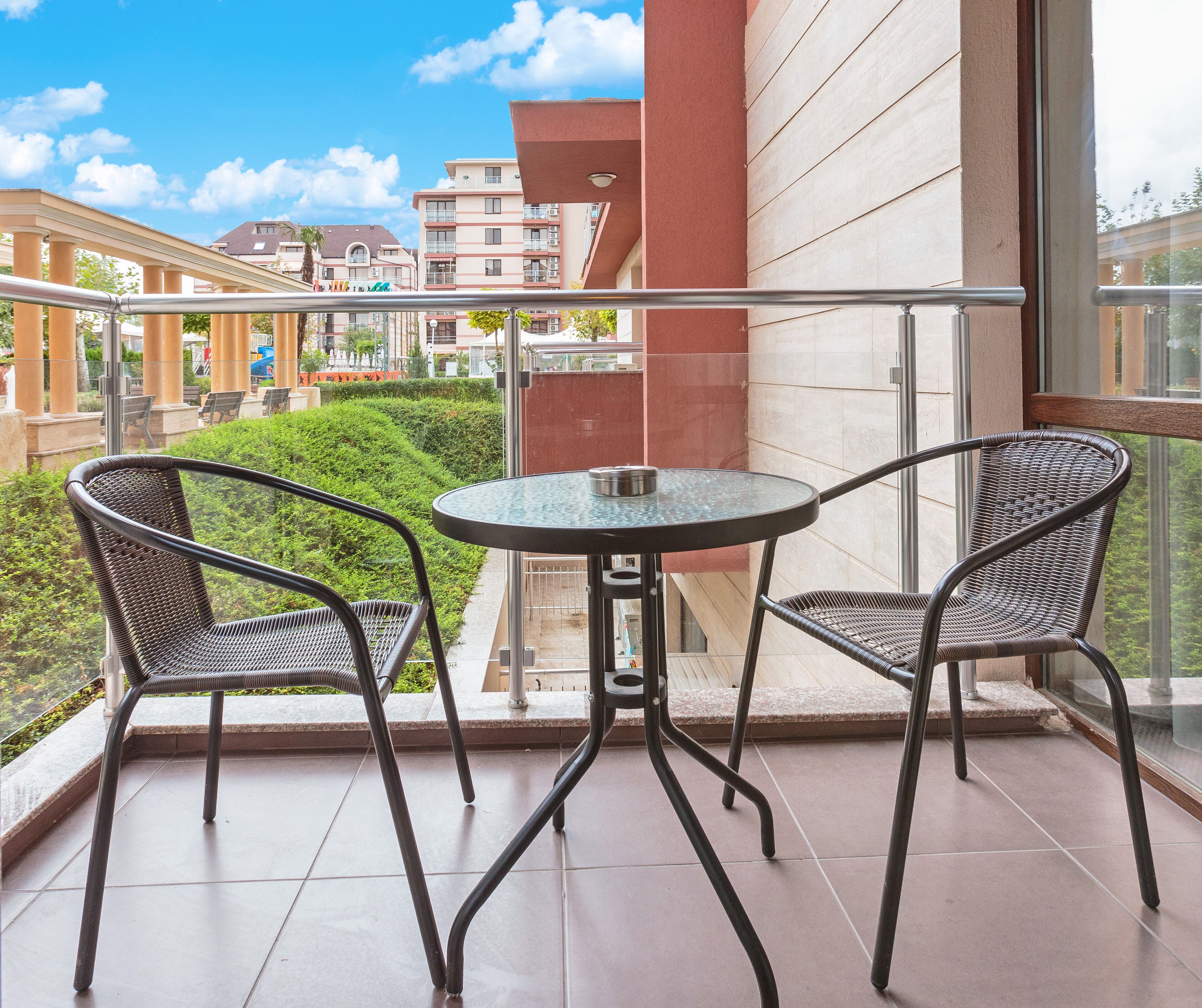 A balcony with two wicker chairs and a glass table, overlooking a landscaped garden and buildings.