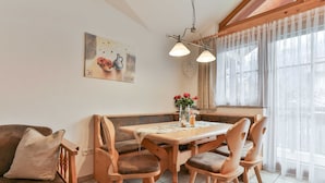 A dining area with a wooden table and chairs, a bench, a painting on the wall, and a window with sheer curtains.