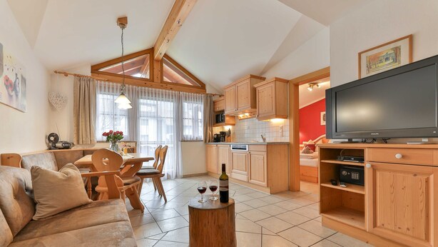 A living room with a sofa, a coffee table with a bottle and glasses, a dining area with chairs, a kitchen with wooden cabinets and a TV.