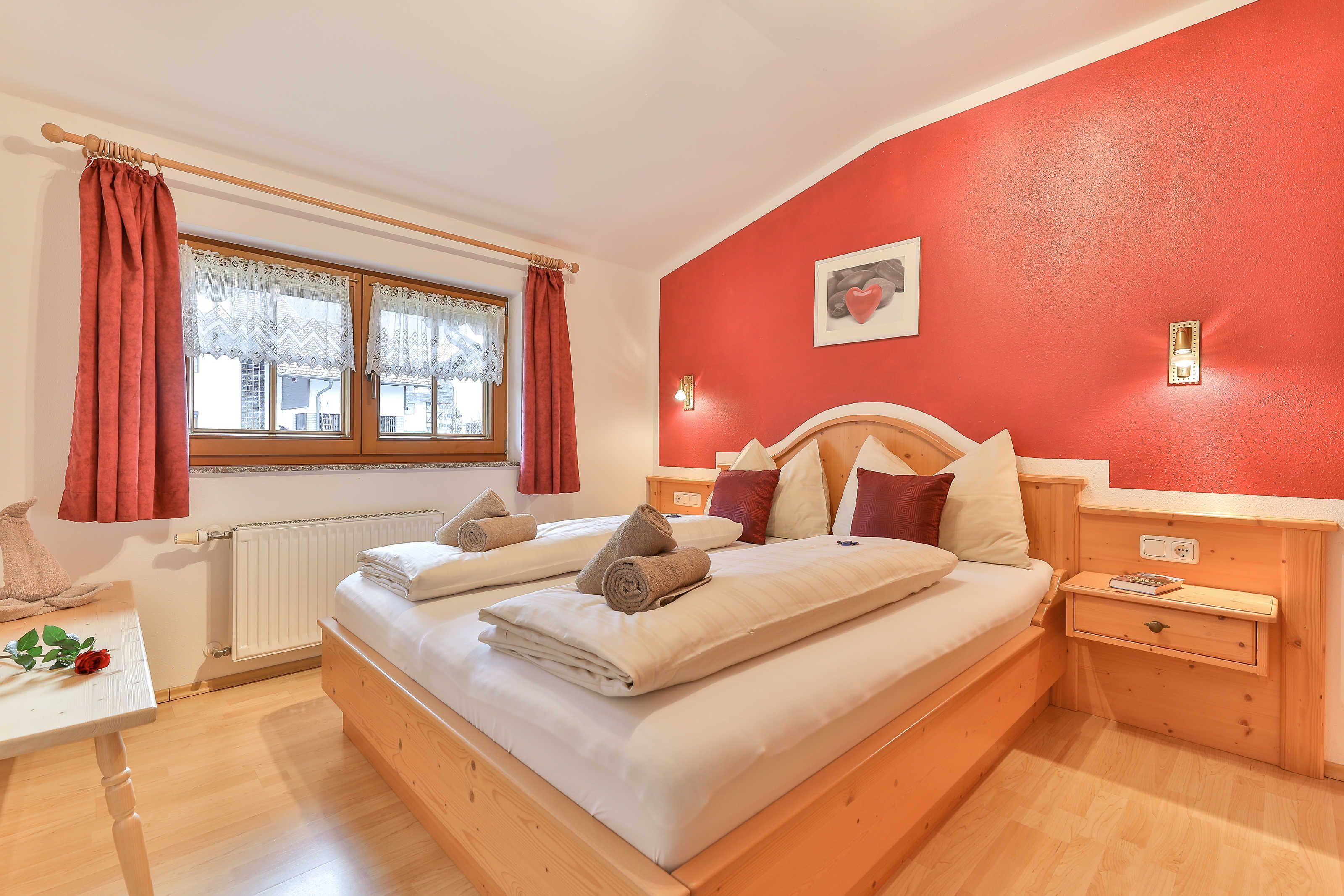 A bedroom with a bed, a nightstand, a table with a rose, and a window with curtains.