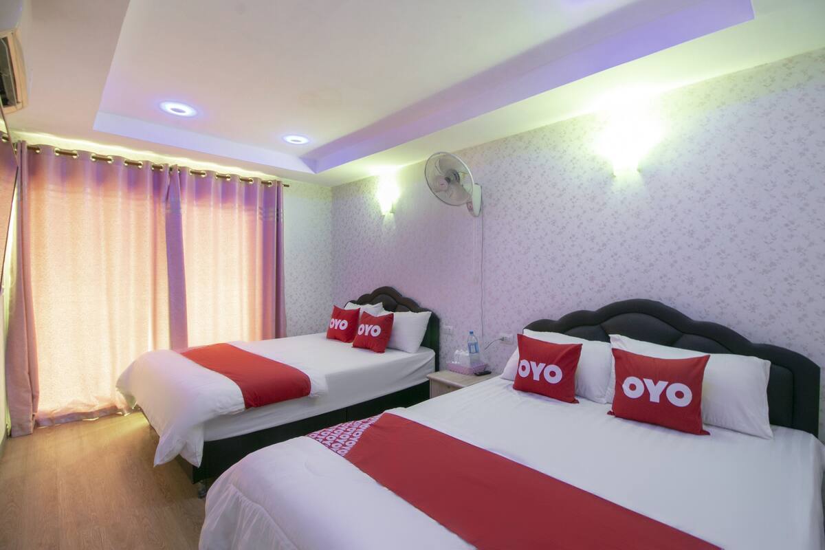 A hotel room with two beds, each with red and white bedding and pillows with the word "OYO". There is a ceiling fan and a wall-mounted light fixture.