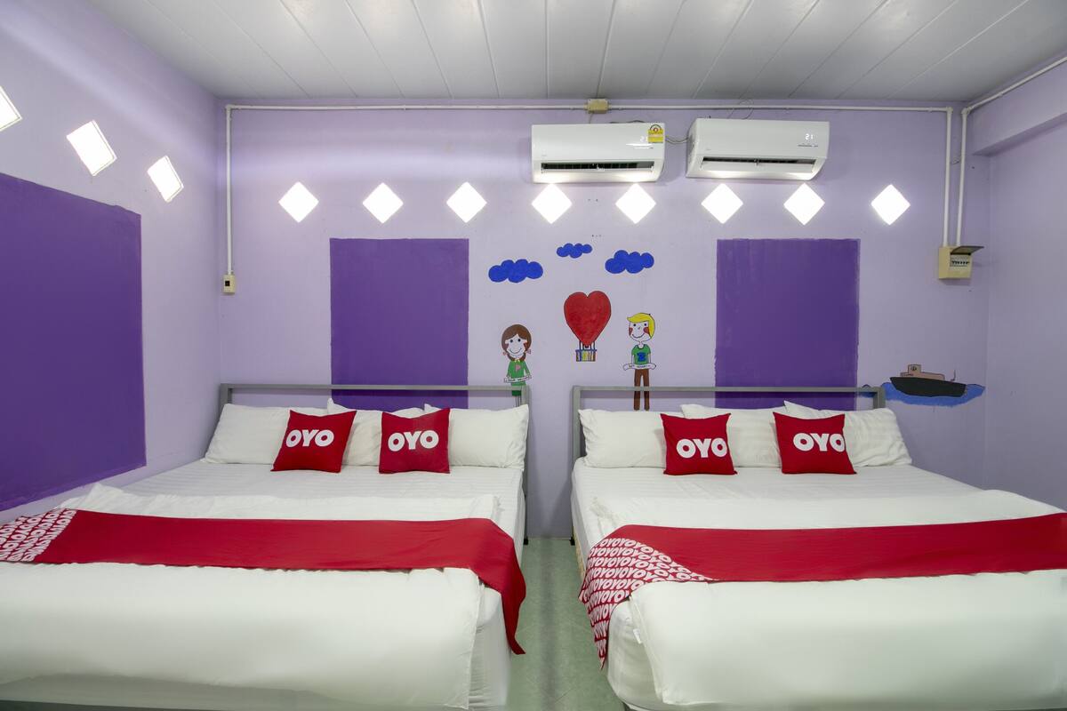 A room with two beds, each with white bedding and red pillows labeled "OYO". There are air conditioning units on the wall, and the room has purple walls and a green floor.