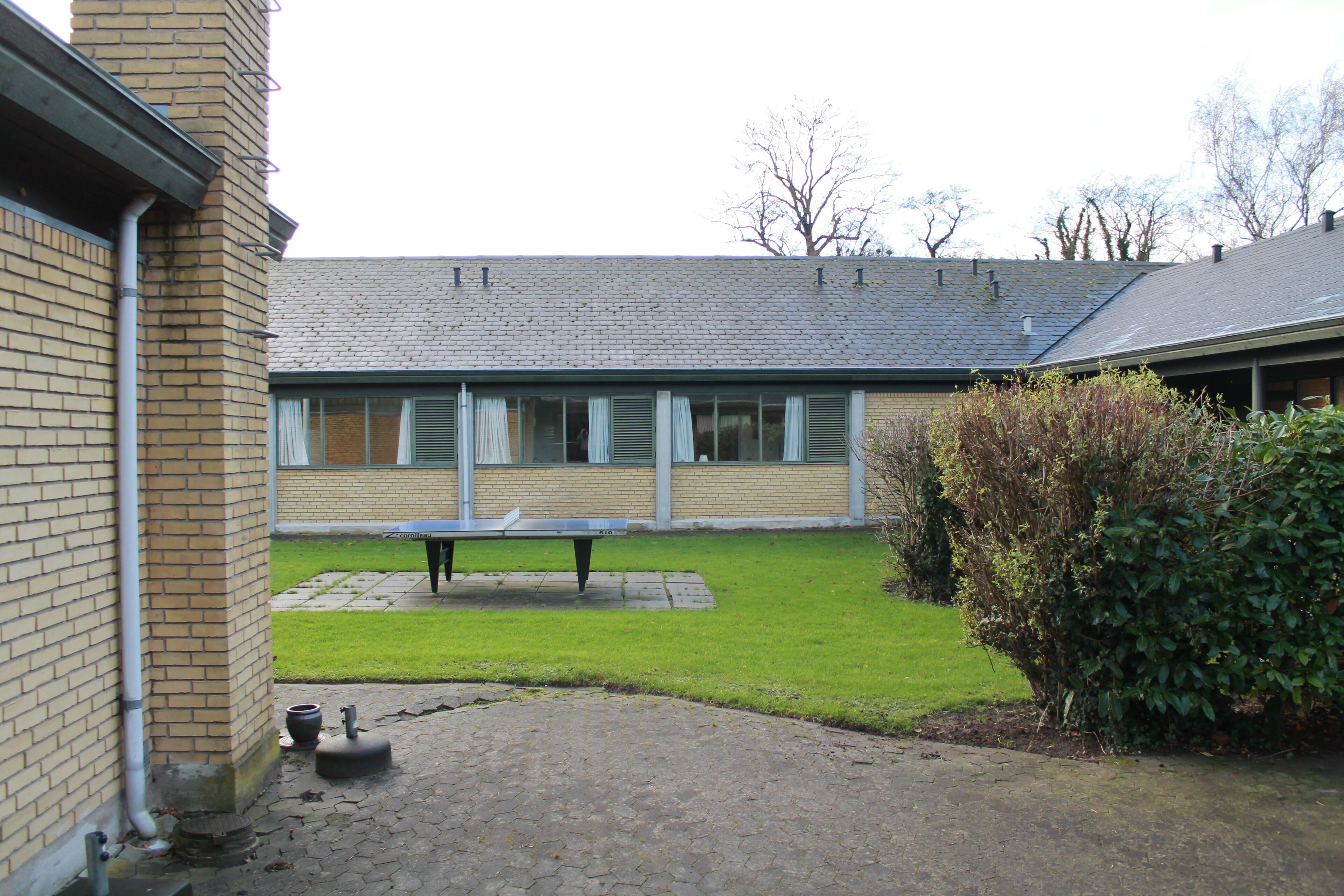 A courtyard with a brick building, a paved area, a bench, and a hedge.