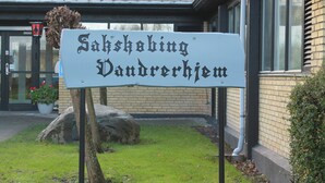 A sign for "Saksholing Vandrerhjem" in front of a building with a brick facade and large windows. - Danhostel Sakskøbing (Sakskobing)