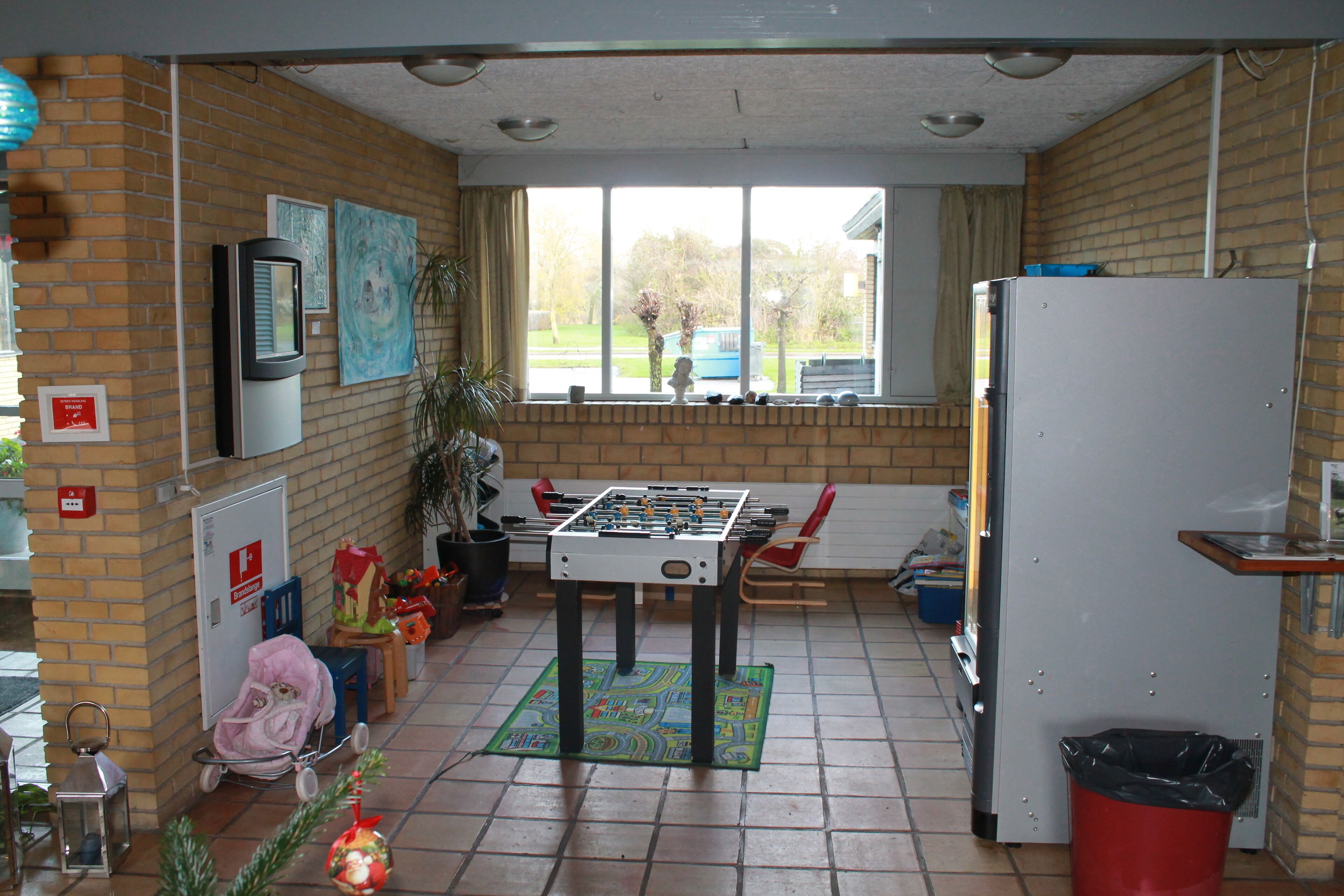 A room with a foosball table, a refrigerator, a trash bin, and a window with a view of the outside.