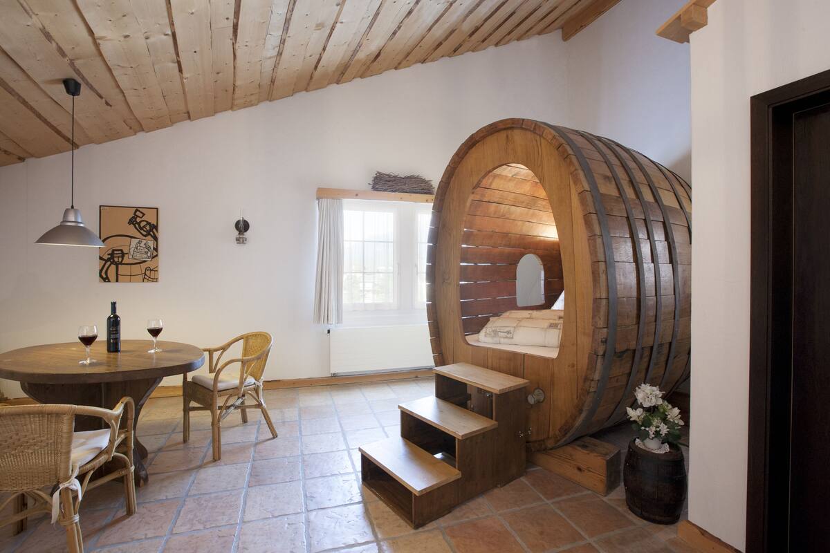 A rustic room with a wooden barrel structure, a round table with chairs, and a small staircase leading to a loft area.