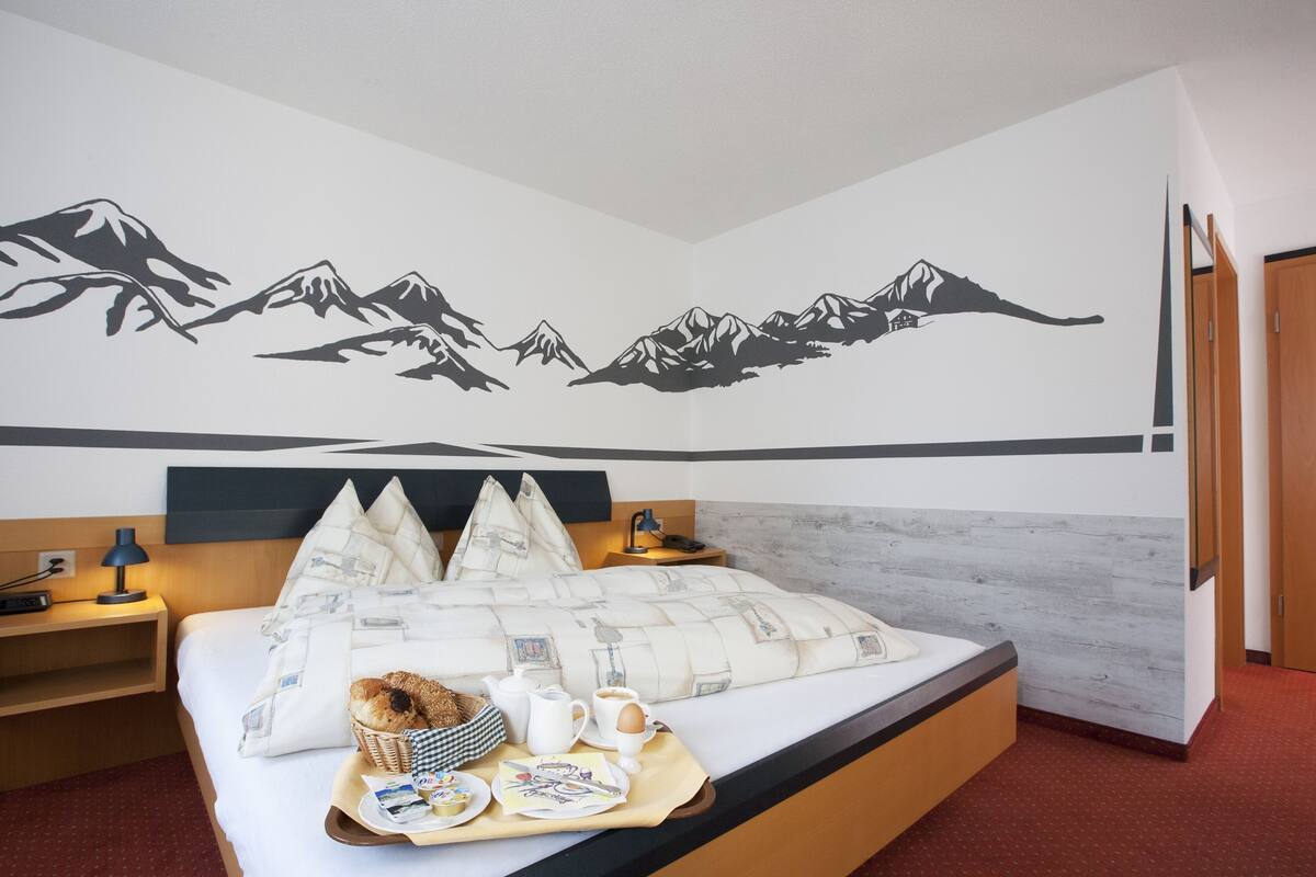 A hotel room with a bed, a wall mural of mountains, a bedside table with a lamp, and a breakfast tray on the bed.